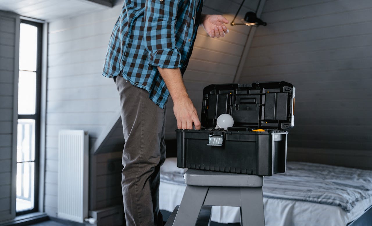 Person in plaid shirt fixing a light bulb using a toolbox in a rustic attic interior.